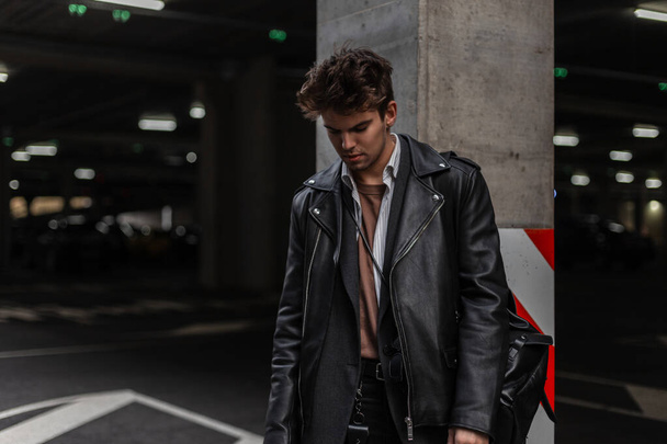 Knappe trendy jongeman model in de jeugd oversized modieuze zwarte leren jas met een stijlvolle kapsel staat en kijkt naar beneden in de buurt van een grijze kolom op een parkeerplaats in de stad. Hipster Amerikaanse moderne man - Foto, afbeelding