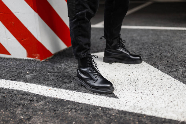 Close-up of fashionable leather seasonal boots on men's legs. Young guy in vintage jeans in trendy shoes stands on the asphalt near a pillar with red-white lines. Stylish casual youth shoes for men. - Фото, изображение