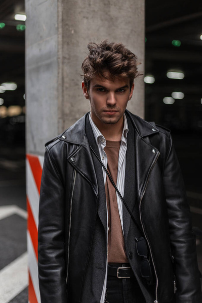 Stylish portrait of a young attractive man hipster with a fashionable hairstyle in trendy black clothes near a pillar on city parking in the street. European handsome guy fashion model posing outdoors - Φωτογραφία, εικόνα