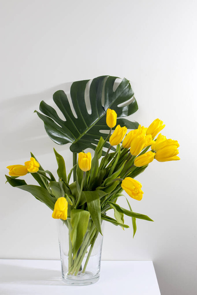 Hoja de monstera en jarra y ramo de tulipanes amarillos está sobre la mesa opuesta pared blanca
 - Foto, Imagen