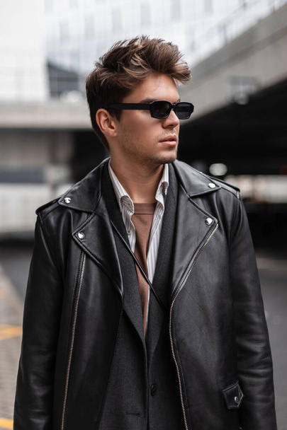 Fresh stylish portrait of a young man hipster in stylish sunglasses with a fashionable hairstyle in trendy black clothes in the street. American handsome guy model posing outdoors. Trendy menswear. - Photo, Image