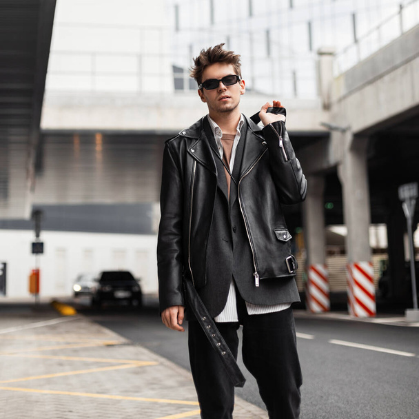 Amerikanische Mode junger Mann Modell in einem stilvollen übergroßen schwarzen Lederjacke mit dunkler Sonnenbrille in Vintage-Jeans geht auf der Straße in der Stadt.Trendy schöner Hipster-Typ steht in der Nähe der Straße im Freien. - Foto, Bild