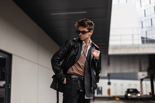 Komea eurooppalainen nuori mies muodissa nuorten ylisuuret vaatteet trendikkäällä kampauksella lepää lähellä modernia rakennusta kaupungissa kevätpäivänä. Moderni kaveri hipster tyylikäs aurinkolasit ulkona
. - Valokuva, kuva