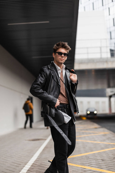 Handsome American young man in a stylish oversized leather black jacket in stylish sunglasses in vintage jeans walks down the street in the city.Attractive trendy hipster guy stands near gray building - Fotoğraf, Görsel
