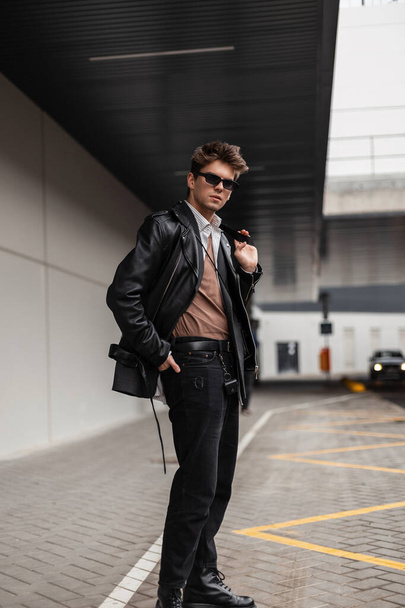 Ein junger europäischer Hipster mit cooler Sonnenbrille, modisch schwarzer Lederjacke, klassischem Hemd in Jeans und Frisur spaziert in der Nähe eines grauen Gebäudes auf der Straße. Attraktiver Trendtyp in der Stadt - Foto, Bild
