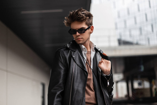 Stilvolles Porträt eines jungen Mannes Hipster mit Sonnenbrille und modischer Frisur in trendiger schwarzer Kleidung in der Nähe von grauen Gebäude in der Stadt auf der Straße. Europäische gutaussehende Kerl Mode Modell posiert im Freien - Foto, Bild
