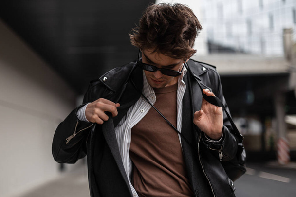 Elegante hombre hipster joven fresco en gafas de sol oscuras de moda en una camisa en una chaqueta de moda vintage posando en la ciudad en un día de primavera. Un tipo atractivo con ropa juvenil en la calle. Estilo moderno americano
 - Foto, Imagen