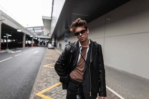 Jonge trendy knappe man hipster in zonnebril met kapsel in een vintage shirt in een stijlvol zwart leren jasje in modieuze jeans loopt op straat in de stad. Aantrekkelijk man model buiten. - Foto, afbeelding