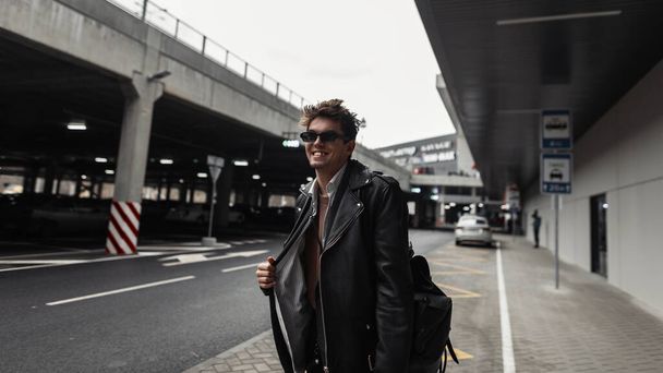 Happy young man hipster with funny smile in a fashionable leather black jacket with a trendy hairstyle stands and smiling on a street in the city. Joyful stylish guy model in sunglasses outdoors.  - Photo, Image