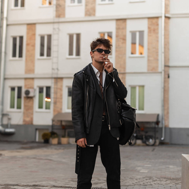 Tyylikäs muotokuva nuori mies savuke muoti aurinkolasit viileä nahka musta takki trendikäs reppu kaupungin kadulla. Kaupunkien hipster kaveri kulumista amerikkalaiseen tyyliin tupakoi ulkona
 - Valokuva, kuva