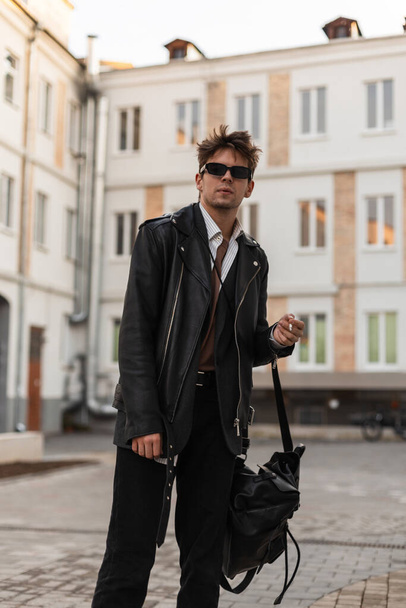 Trendy young man in youth oversized fashionable black leather jacket with hairstyle with backpack stands with cigarette near modern building in the city. Guy hipster in sunglasses smokes outdoors. - Photo, image