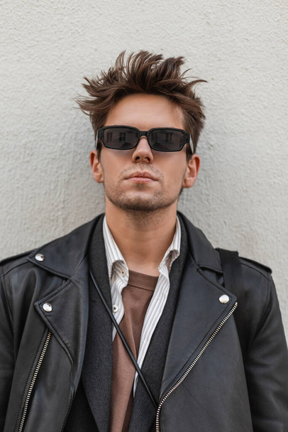 Fashionable portrait young man with serious face in a vintage dark sunglasses in a trendy leather black jacket with a stylish hairstyle near the wall outdoors. Handsome modern hipster guy in the city. - Foto, Imagen