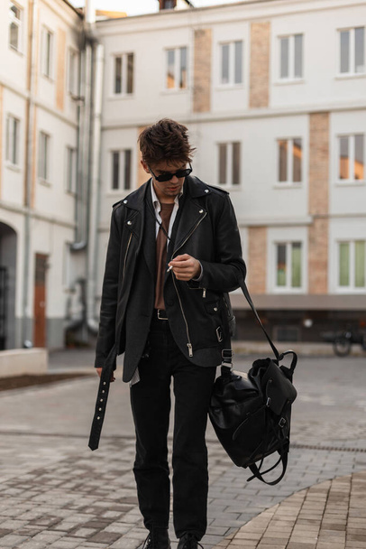 Young handsome man hipster in sunglasses in shirt in a stylish black leather jacket with backpack smokes near the buildings in the city. Cool guy fashion model with cigarette outdoors. American style. - Фото, зображення