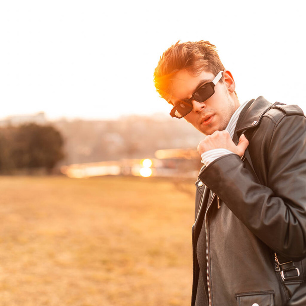 Divatos modell fiatalember elegáns napszemüveg divatos bőr fekete kabát ing pihen a szabadban a háttérben csodálatos naplemente. Vonzó hipszter srác élvezi a narancssárga napfény a természetben. - Fotó, kép