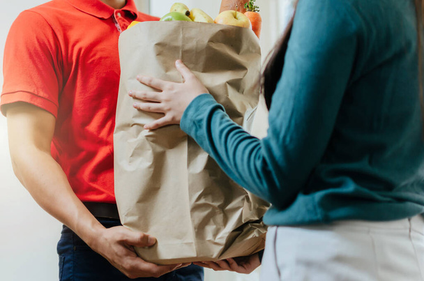 smart food delivery service man in red uniform handing fresh food to recipient and young woman customer receiving order from courier at home, express delivery, food delivery, online shopping concept - Foto, Imagen