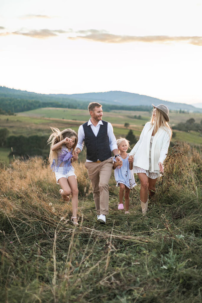 Joyful Caucasian family with two children enjoying summer walk in countryside field in mountains. Family in stylish boho casual clothes walking in the field holding hands - Φωτογραφία, εικόνα