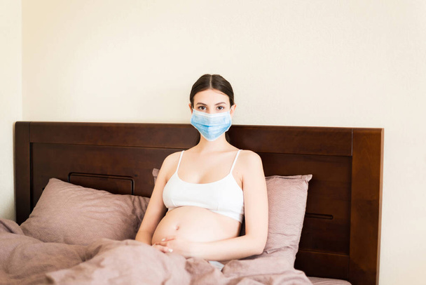 Egy fiatal terhes nő orvosi maszkban ágyban marad a koronavírus miatt. Karantén idő. Egészségügyi koncepció. - Fotó, kép