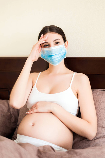 Terhes nő védő orvosi maszk van coronavirus és marad ágyban szenved fejfájás. Megelőzni a magzat fertőzését. Globális járvány. A karantén koncepciójának fenntartása. - Fotó, kép