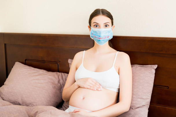 Shocked pregnant woman is wearing protective medical mask with coronavirus word. Mother is staying in bed because of respiratory disease. Global pandemic concept. Maintain the quarantine concept. - Foto, imagen