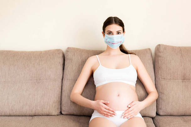 Нездоровая беременная женщина в защитной медицинской маске сидит дома на диване. Эпидемия коронавируса. Дыхательные заболевания
. - Фото, изображение