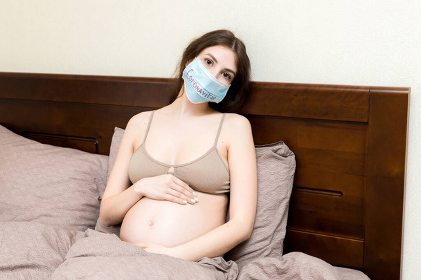 Pregnant woman in protective medical mask with coronavirus word is staying in bed because of feeling sick. Global epidemic concept. Quarantine concept. - Photo, Image