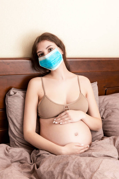 Hamile kadın koruyucu tıp maskesi kötü hissettiriyor ve bu yüzden yatakta kalıyor. Coronavirüs salgını. Genel Salgın. - Fotoğraf, Görsel