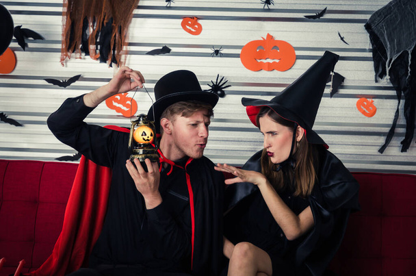 Halloween. couple young caucasian man and woman in vampires and witch clothing holding pumpkin head lantern and sitting on sofa in halloween party at home, holiday and halloween festival concept - Zdjęcie, obraz