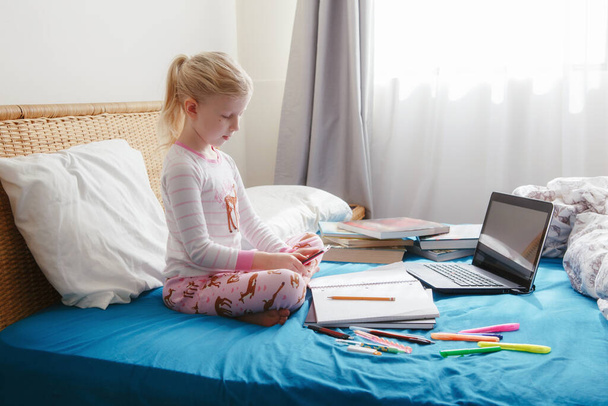 Caucasian girl child sitting in bed and learning online on laptop Internet. Virtual class lesson on video during self isolation at home. Distant remote video education. Modern school study for kids. - Foto, immagini