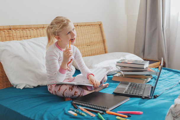 Caucasian girl child sitting in bed and learning online on laptop Internet. Virtual class lesson on video during self isolation at home. Distant remote video education. Modern school study for kids. - 写真・画像