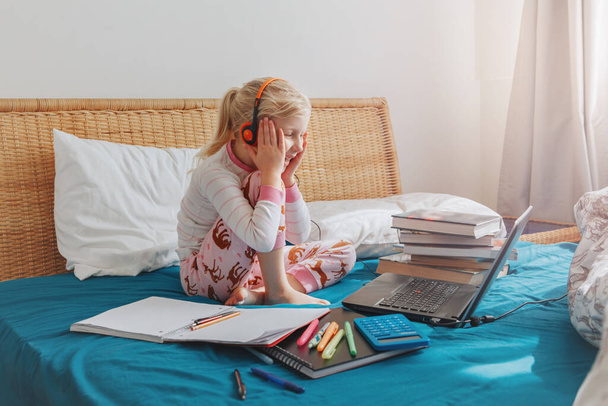 Biała dziewczyna dziecko siedzi w łóżku i uczy się online na laptopie Internet. Lekcja wirtualnej klasy na wideo podczas samodzielnej izolacji w domu. Oddalona edukacja wideo. Nowoczesne badania szkolne dla dzieci. - Zdjęcie, obraz