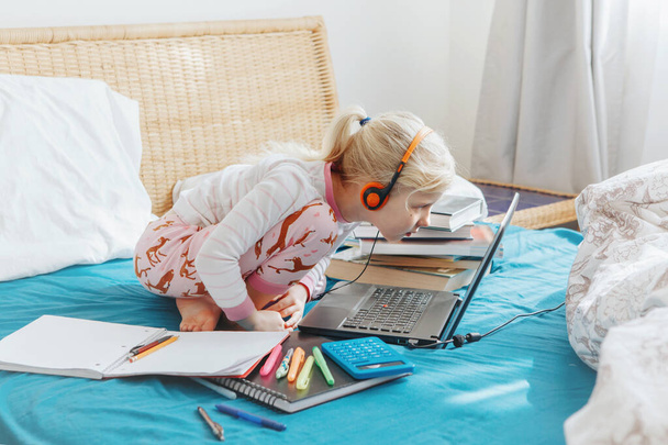 Kaukasisch meisje kind zitten in bed en leren online op laptop Internet. Virtuele les op video tijdens zelfisolatie thuis. Afstandsonderwijs op afstand. Moderne school studie voor kinderen. - Foto, afbeelding