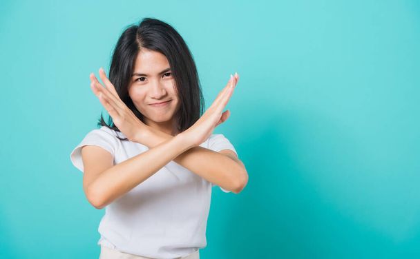 Portrait happy Asian beautiful young woman unhappy or confident standing wear white t-shirt, She holding two crossing arms say no X sign, studio shot on blue background with copy space for text - Zdjęcie, obraz