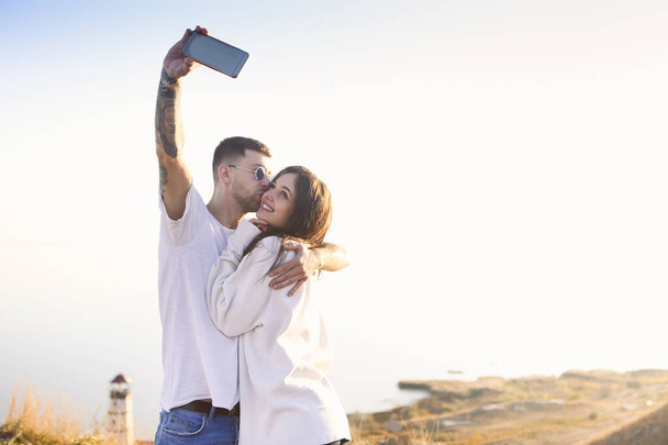 Positive young romantic couple in casual clothes hugging and kissing while taking selfie with smartphone on beach with lighthouse in background in sunny summer day - Photo, Image