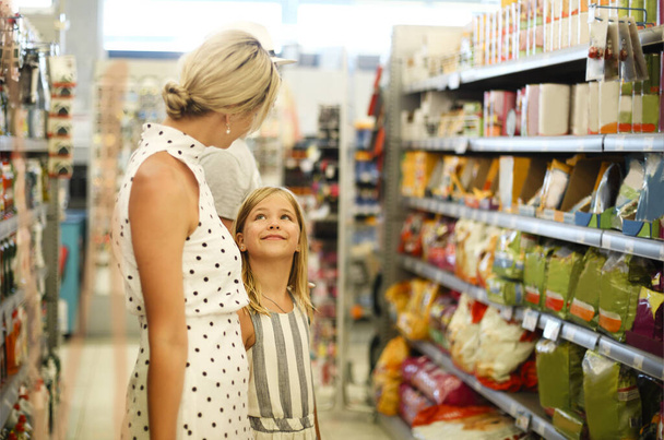 Uśmiechnięta młoda blond matka z córeczką wybierająca produkty stojąc we współczesnym supermarkecie. Koncepcja zakupów rodzinnych - Zdjęcie, obraz