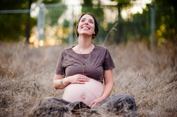 Ευτυχισμένη νεαρή έγκυος γυναίκα χαμογελά καθισμένη στα χωράφια. - Φωτογραφία, εικόνα