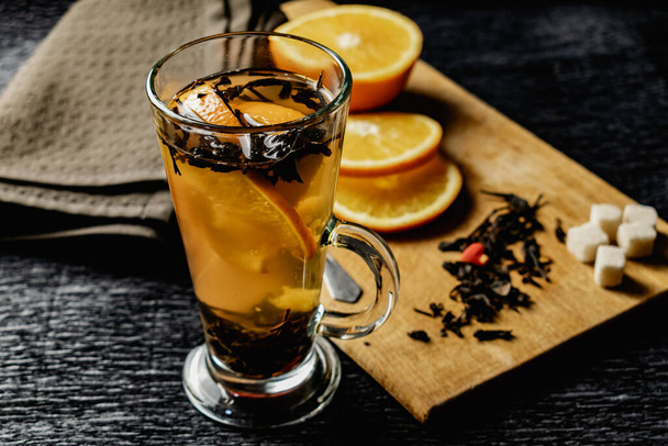 Citrus drink, tea with petals in a transparent glass mug with slices of sweet orange and refined sugar. Slicing fruit on a wooden Board with a brown napkin in a cozy dark background in a low key - Photo, Image