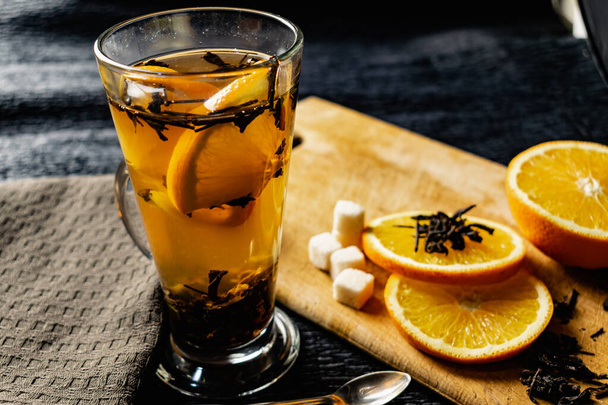 Citrus drink, tea with petals in a transparent glass mug with slices of sweet orange and refined sugar. Slicing fruit on a wooden Board with a brown napkin in a cozy dark background in a low key - Photo, Image