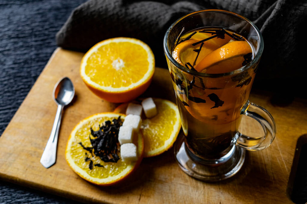 Citrusital, tea szirmokkal egy átlátszó üvegbögrében, édes narancs szeletekkel és finomított cukorral. Gyümölcs szeletelése egy fa fórumon barna szalvétával, hangulatos sötét háttérrel, alacsony kulccsal - Fotó, kép