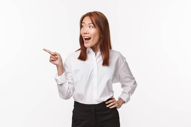 retrato de animado e divertido elegante jovem asiático mulher no branco escritório camisa, apontando olhando dedo esquerdo e sorrindo emocionado como ver algo interessante, branco fundo
 - Foto, Imagem