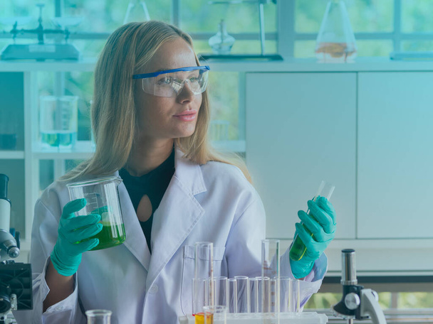 Pretty Caucasian scientist woman holding glass beaker and test tube filled with green liquid. - Photo, Image