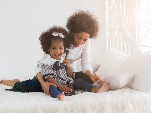 Cute little african girl playing with vintage old dial phone on the white bed with mom. Bonding time, educational playtime. - Photo, Image