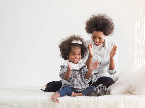 Dark skinned mom and cute little daughter clapping their hands on the white bed in the bedroom. - Photo, Image