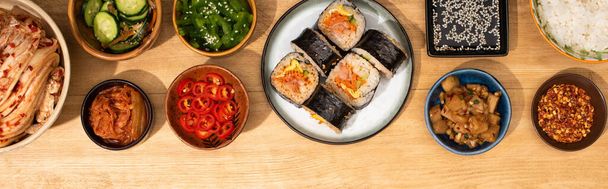 panoramic shot of bowls with korean side dishes near gimbap on plate  - Photo, Image