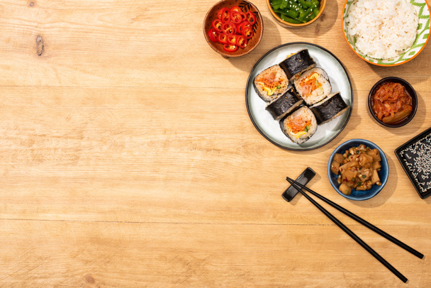 vue sur le dessus des bols avec de savoureux plats d'accompagnement coréens près des baguettes et gimbap sur la surface en bois
  - Photo, image