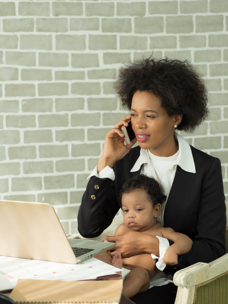 Imagen vertical, madre africana trabajando y usando el teléfono móvil mientras cuida al niño sentado en el regazo. Madre soltera, mujer de negocios trabajar en casa. - Foto, Imagen