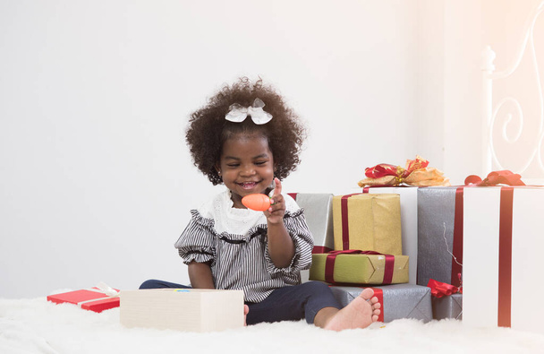 Schattig Afrikaans meisje zittend tussen cadeautjes of geschenkdozen en één geopende doos. Schattig meisje met speelgoed wortel met geluk. Verjaardag, Nieuwjaar, Kerstconcept. - Foto, afbeelding