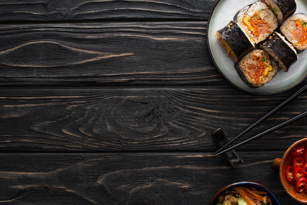 верхний вид палочек для еды рядом с тарелкой с корейским джимбапом и вкусными гарнирами на деревянной поверхности
  - Фото, изображение