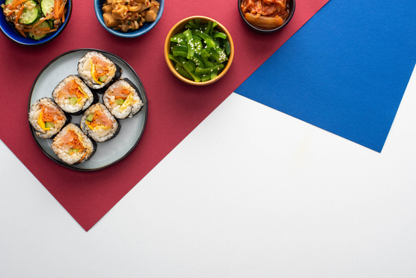vrchní pohled na talíř s čerstvým gimbapem v blízkosti korejských bočních jídel na bílém, modrém a karmínovém  - Fotografie, Obrázek