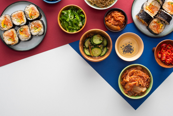 vista superior de placas com gimbap fresco perto de pratos laterais coreanos em branco, azul e carmesim
  - Foto, Imagem