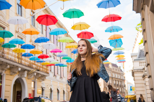 Beautiful girl walking under umbrellas in the city. Cute teenage girl in black dress posing against a background of multicolored umbrellas alley. Spring sunny concept. 8 March. Happy Women's Day. - Zdjęcie, obraz